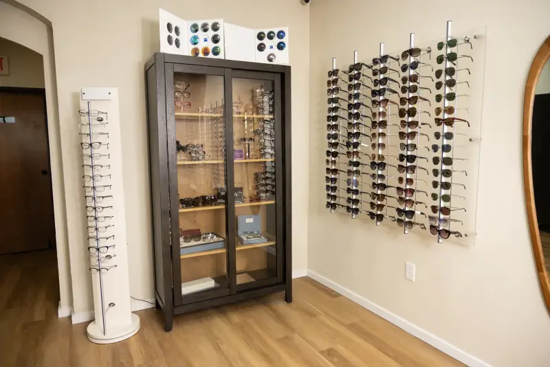 A wall panel of sunglasses beside a tall wooden display cabinet of frames