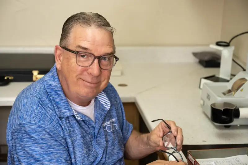 Robert Spurrier at the lab bench holding a pair of eyeglasses beside the edging equipment
